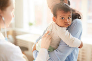 Young mother holding her little child on her hands while female doctor examining him with stethoscope