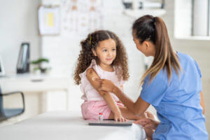 A sweet little girl of Latin decent, sits up straight on an exam table as her nurse places a bandage on her arm after giving her a vaccination. The nurse is wearing blue scrubs and smiling at the young girl to put her at ease.